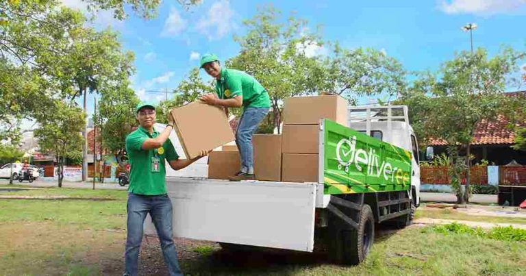 Dua pekerja sedang memuat kotak ke dalam truk pengiriman yang berwarna hijau dengan logo 'Deliveree'